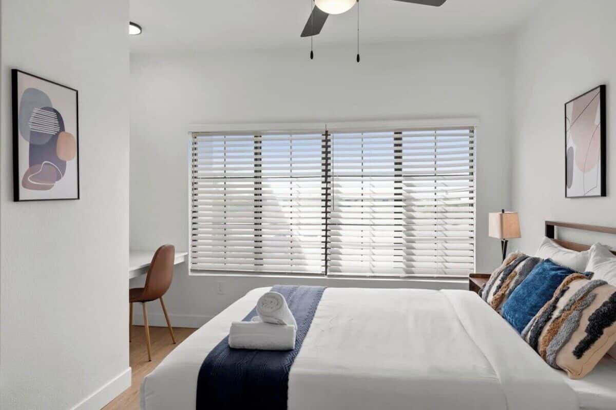 A bedroom with a bed, a desk, and a chair. There are two windows with blinds, a ceiling fan, and wall art.