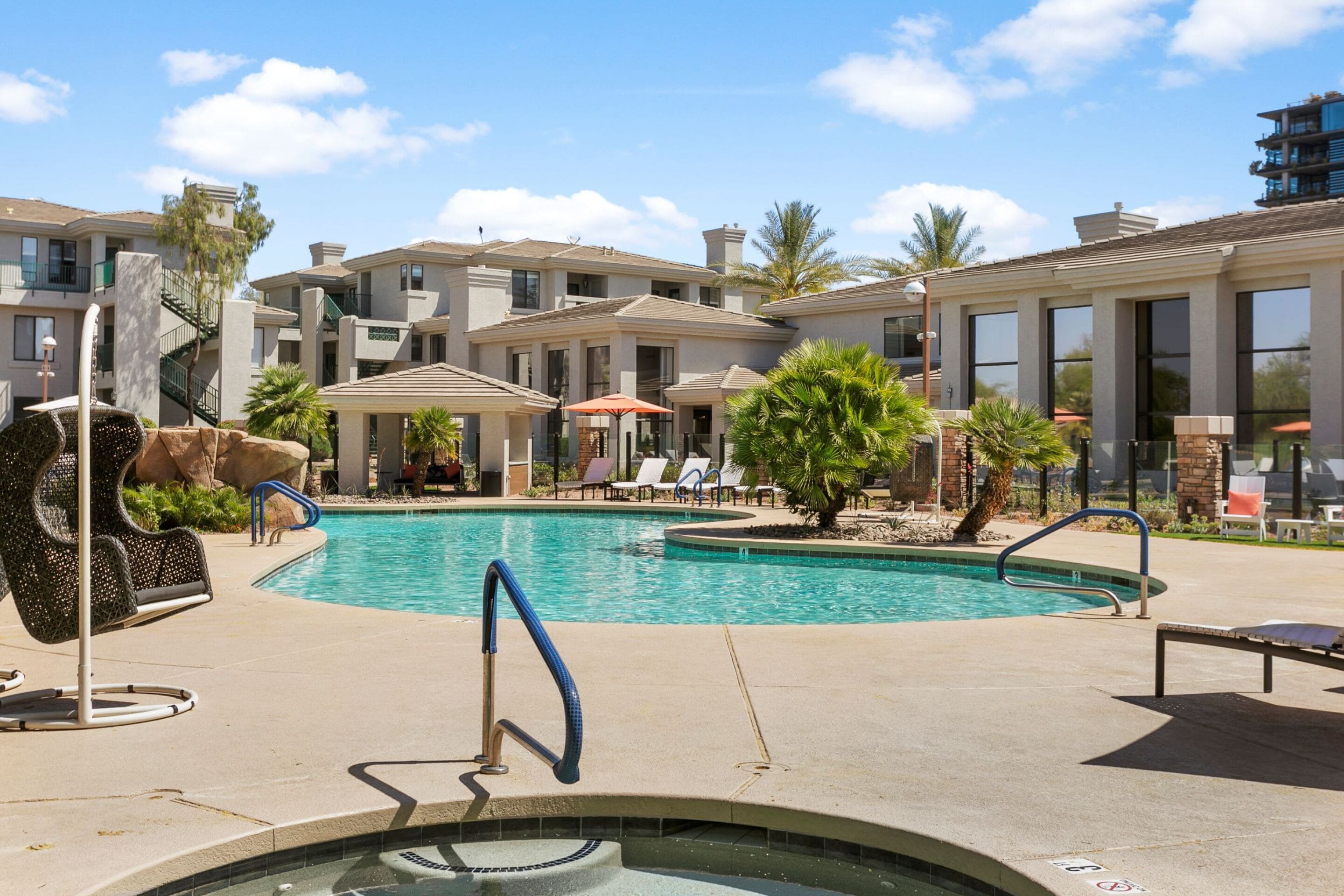 Una piscina con tumbonas y sombrillas, rodeada de edificios con balcones y una palmera.