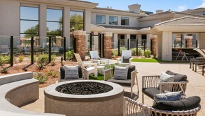 A modern outdoor patio area with seating, a fire pit, and a view of a large house with multiple windows and a covered section. - CozySuites at Kierland Commons (Scottsdale)