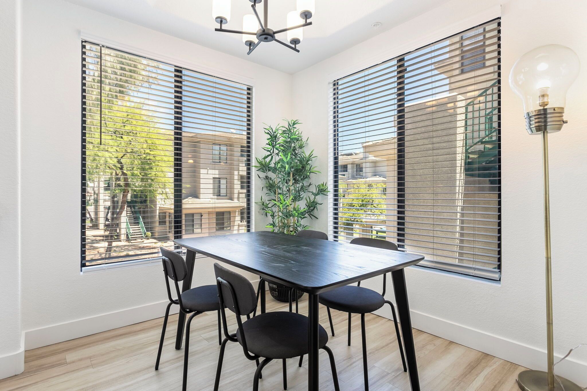 Un área de comedor con una mesa y sillas, ventanas amplias con persianas, una planta en maceta y una lámpara de pie.