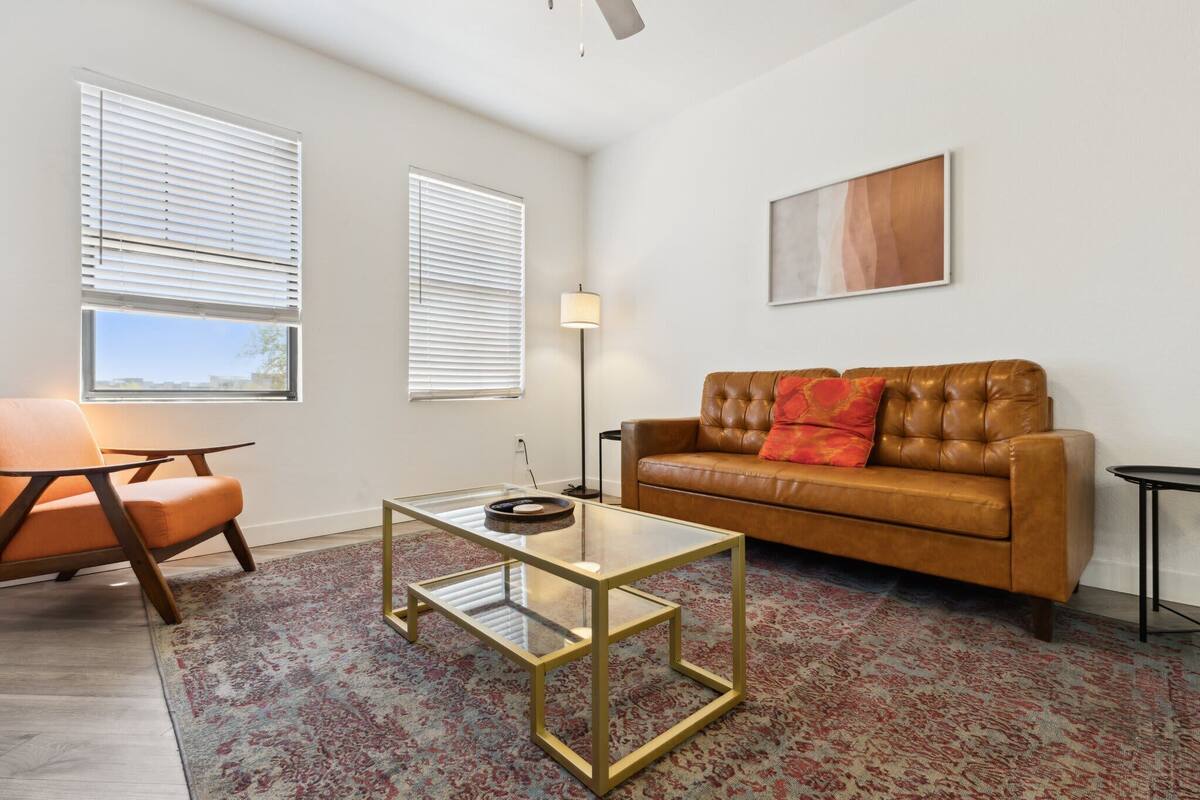 A living room with a brown leather sofa, a glass coffee table, a patterned rug, and two windows with blinds.