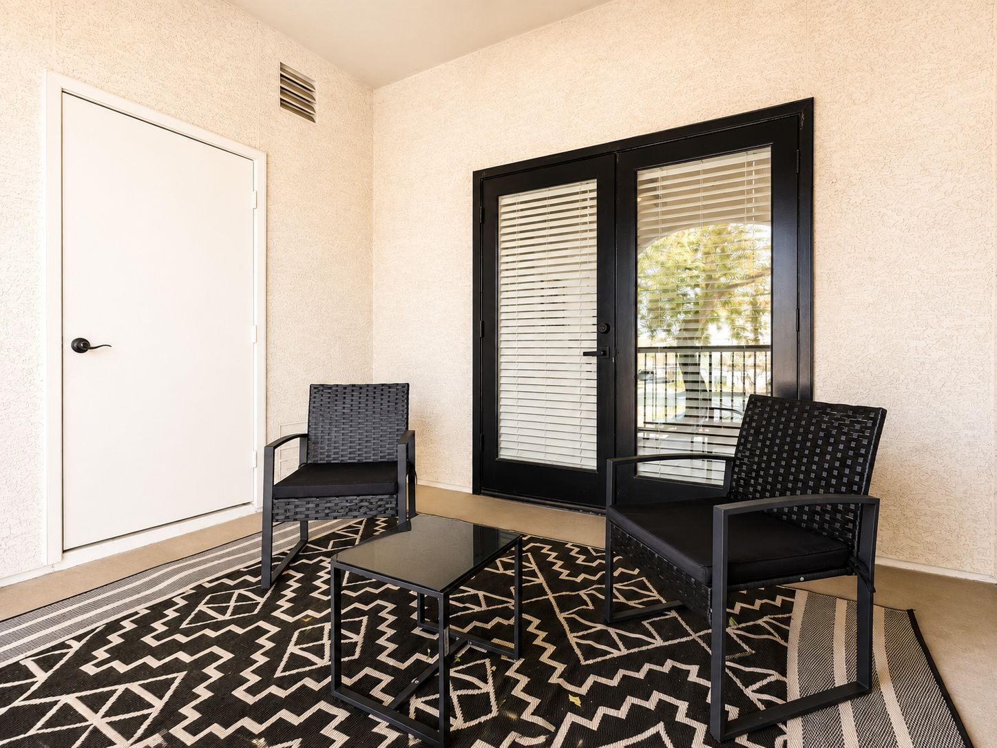 A small balcony with two chairs and a table, a door, and a window with blinds.
