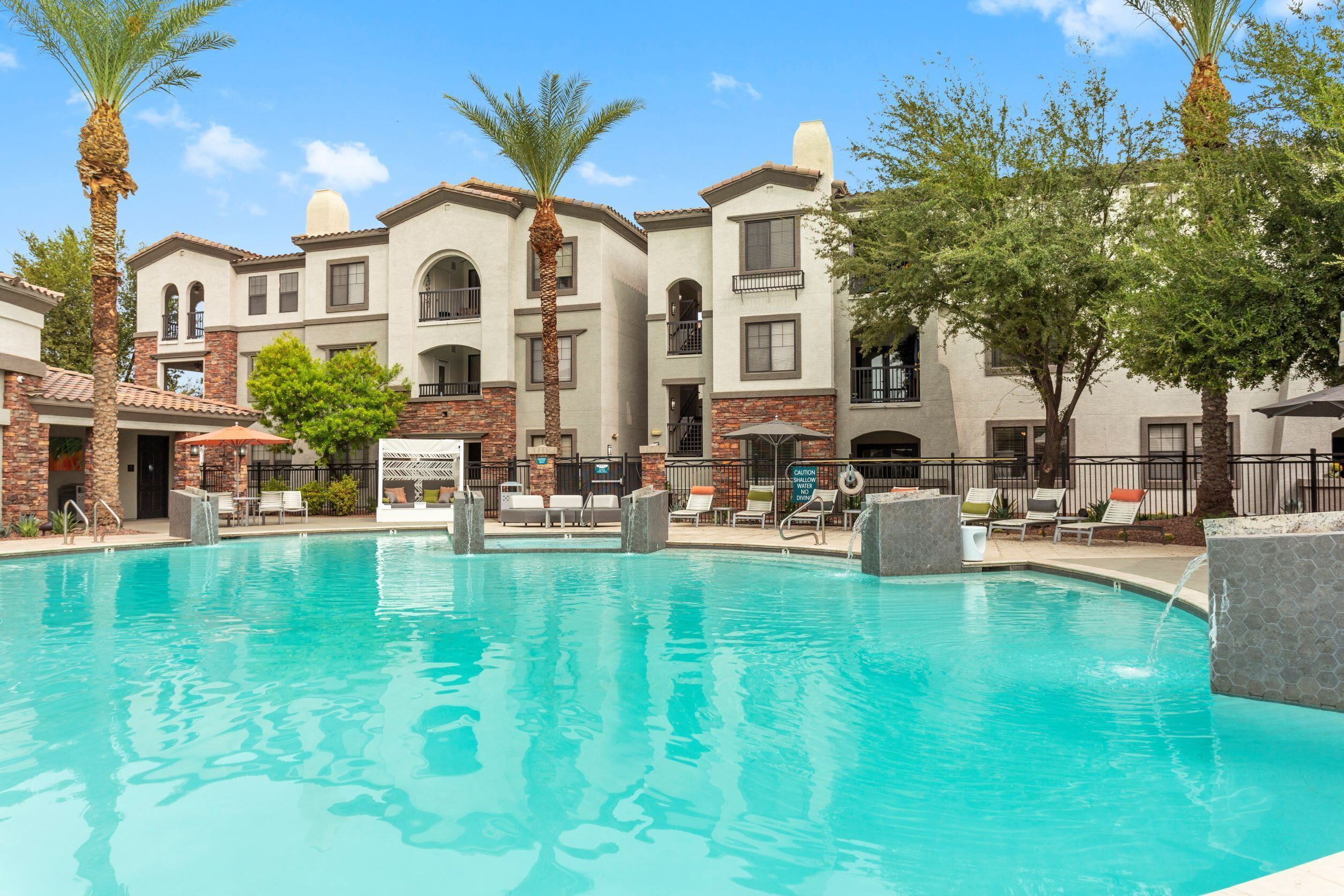A swimming pool with lounge chairs and palm trees, surrounded by multi-story buildings with balconies.