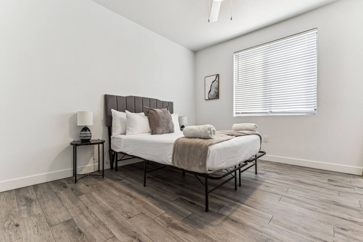 A modern bedroom with a bed, bedside tables, a window with blinds, and a wall-mounted artwork.