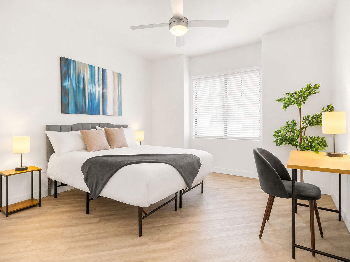 A modern bedroom with a bed, bedside tables, a desk with a chair, a ceiling fan, and a window with blinds.