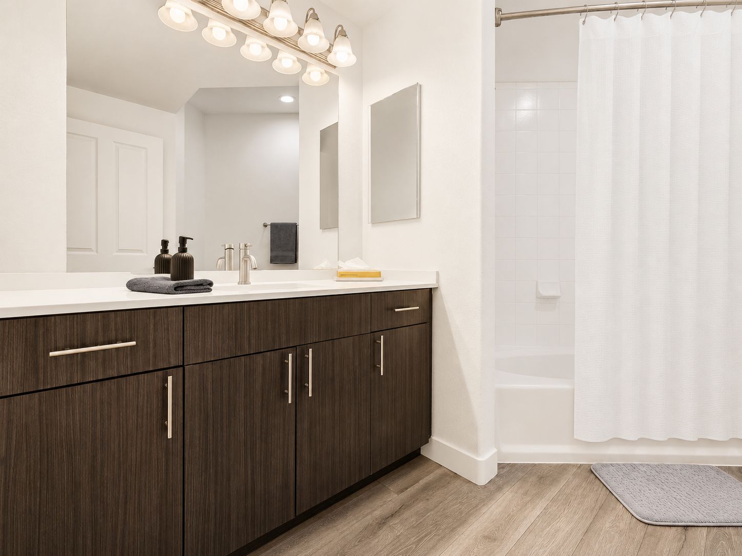 A modern bathroom with a large mirror, a vanity with dark wood cabinets, a white sink, and a shower area with a white curtain.