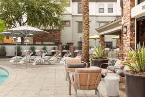 Zona junto a la piscina con sillones de descanso y sombrillas, una palmera y un edificio de fachada de piedra.