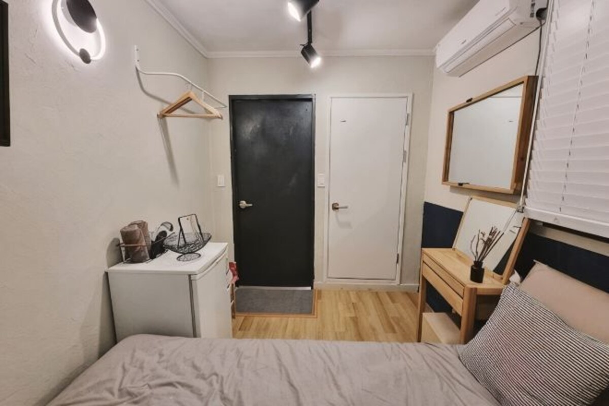 A bedroom with a bed, a desk, a chair, a small table, a mirror, and a door.