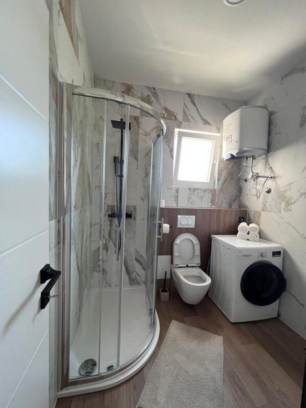A bathroom with a glass shower enclosure, a toilet, a washing machine, and a small window.