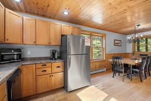 A kitchen with wooden cabinets, a stainless steel refrigerator, a coffee maker, and a dining table with chairs. - Maine Lodge with Fireplace and Pool Table (Bethel)