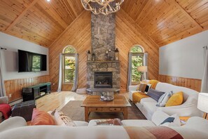 A cozy living room with a stone fireplace, wooden ceiling, and a television mounted on the wall. - Maine Lodge with Fireplace and Pool Table (Bethel)