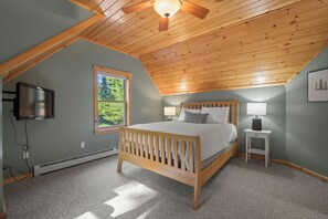 A bedroom with a sloped ceiling, a bed, a nightstand, a lamp, a television, and a window with a view of trees. - Maine Lodge with Fireplace and Pool Table (Bethel)