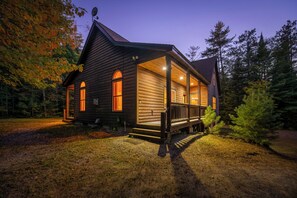 A log cabin with a porch and multiple windows, surrounded by trees. - Maine Lodge with Fireplace and Pool Table (Bethel)