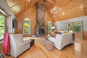 A spacious living room with a stone fireplace, wooden ceiling, and large windows. - Maine Lodge with Fireplace and Pool Table (Bethel)