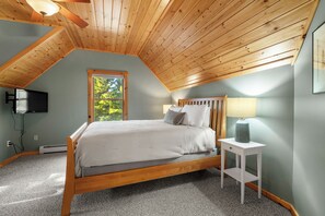 A bedroom with a sloped ceiling, a bed, a nightstand, a lamp, and a TV mounted on the wall. - Maine Lodge with Fireplace and Pool Table (Bethel)