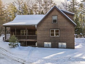 Una cabaña de madera con un porche, una pequeña terraza y un paisaje cubierto de nieve.