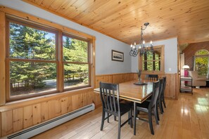A dining area with a wooden table and chairs, large windows with a forest view, a chandelier, and a small kitchen area with a lamp. - Maine Lodge with Fireplace and Pool Table (Bethel)