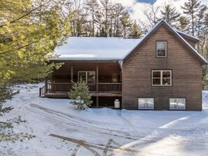 Una cabaña de madera con un porche, rodeada de nieve y árboles.