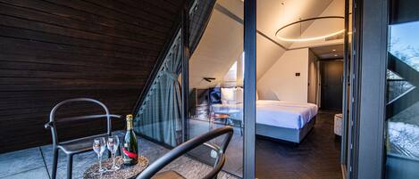 A modern hotel room with a bed, a small table with a bottle and glasses, and a chair.
