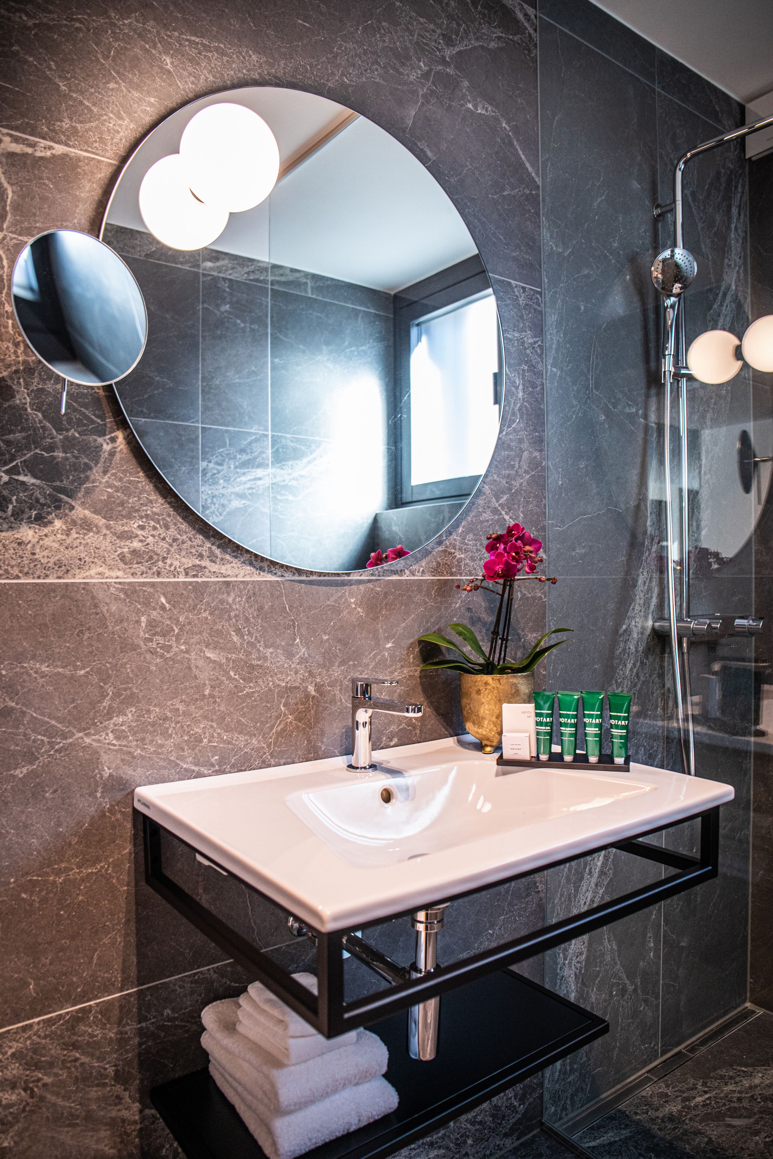Un baño moderno con pared de mármol, un espejo redondo, un lavabo con marco negro y una zona de ducha.