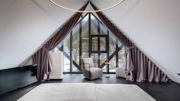 Una habitación moderna de forma triangular con un ventanal grande, una cama, un sillón y un televisor.