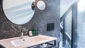 Une salle de bain moderne avec un grand miroir, un lavabo et des toilettes. On trouve des articles de toilette sur le lavabo, et la pièce est dotée d’une grande fenêtre donnant sur l’extérieur.