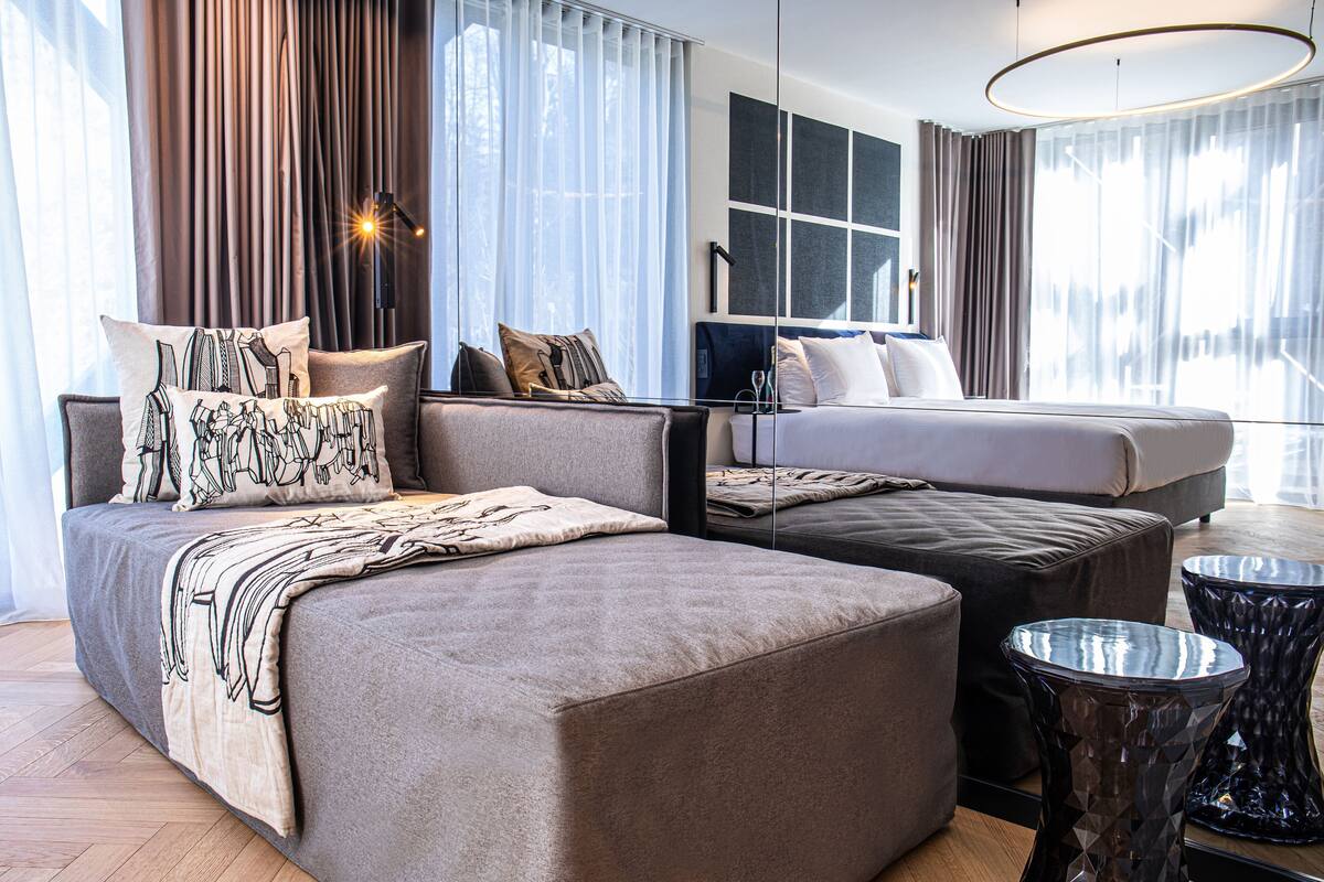 A modern hotel room with a large bed, a sofa, a small table, and a large window with curtains.