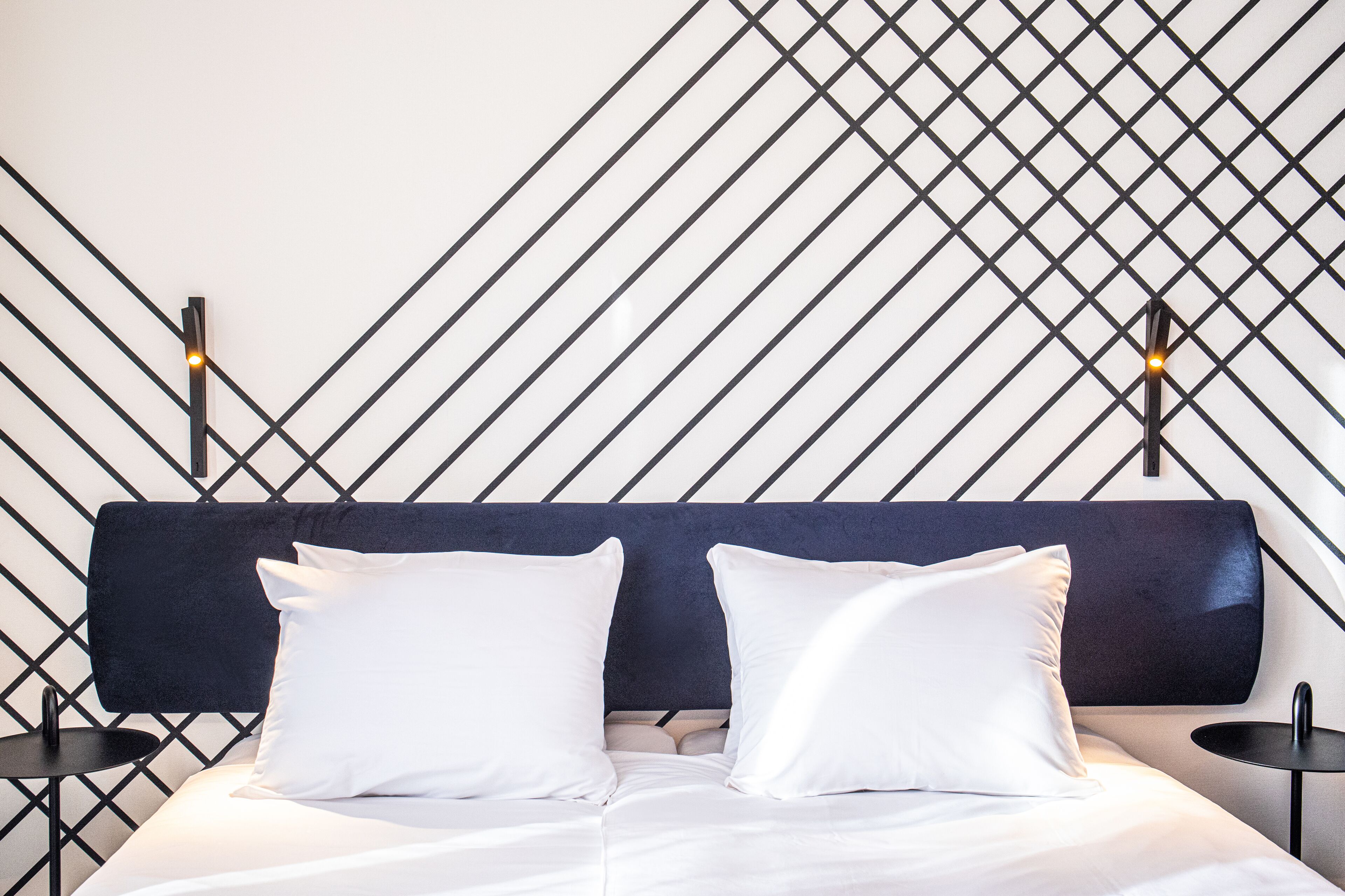 Une chambre d’hôtel moderne avec un mur orné d’un motif géométrique, une tête de lit sombre et deux tables de chevet avec des lampes.