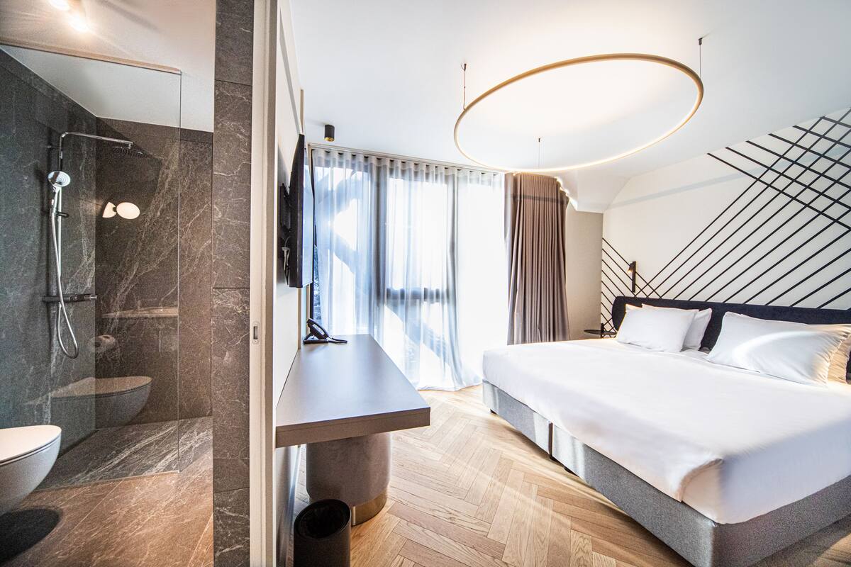 A modern hotel room with a large bed, a shower area, a bedside table, and a view of the outdoors.