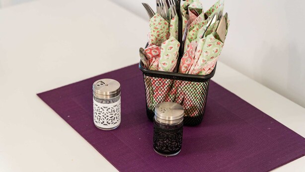 Una mesa con un posavasos morado, un salero de metal y una cesta con servilletas dobladas y cubiertos.