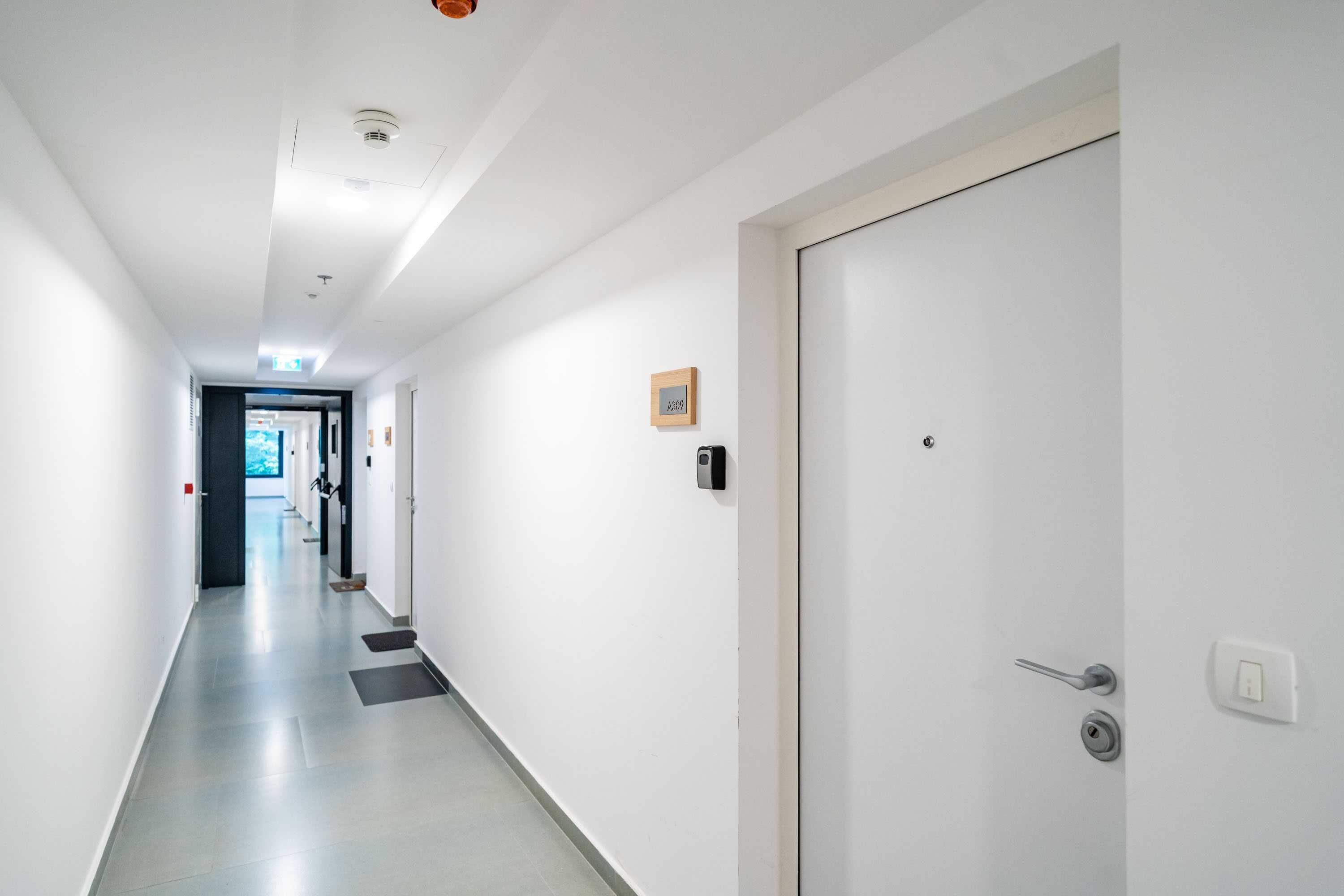 Un pasillo limpio y moderno con una puerta blanca, un pequeño marco de cuadros y un timbre en la pared.