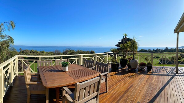 Une terrasse en bois avec une table et des chaises, entourée de plantes en pot, offrant une vue sur la côte.
