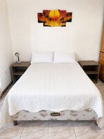 Un dormitorio con una cama grande, ropa de cama blanca, una mesita de noche con lámpara, un armario de madera y un cuadro de atardecer colgado en la pared.