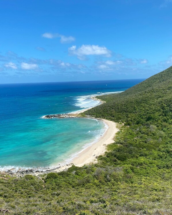 A coastal landscape with a beach, clear turquoise waters, and dense green vegetation.