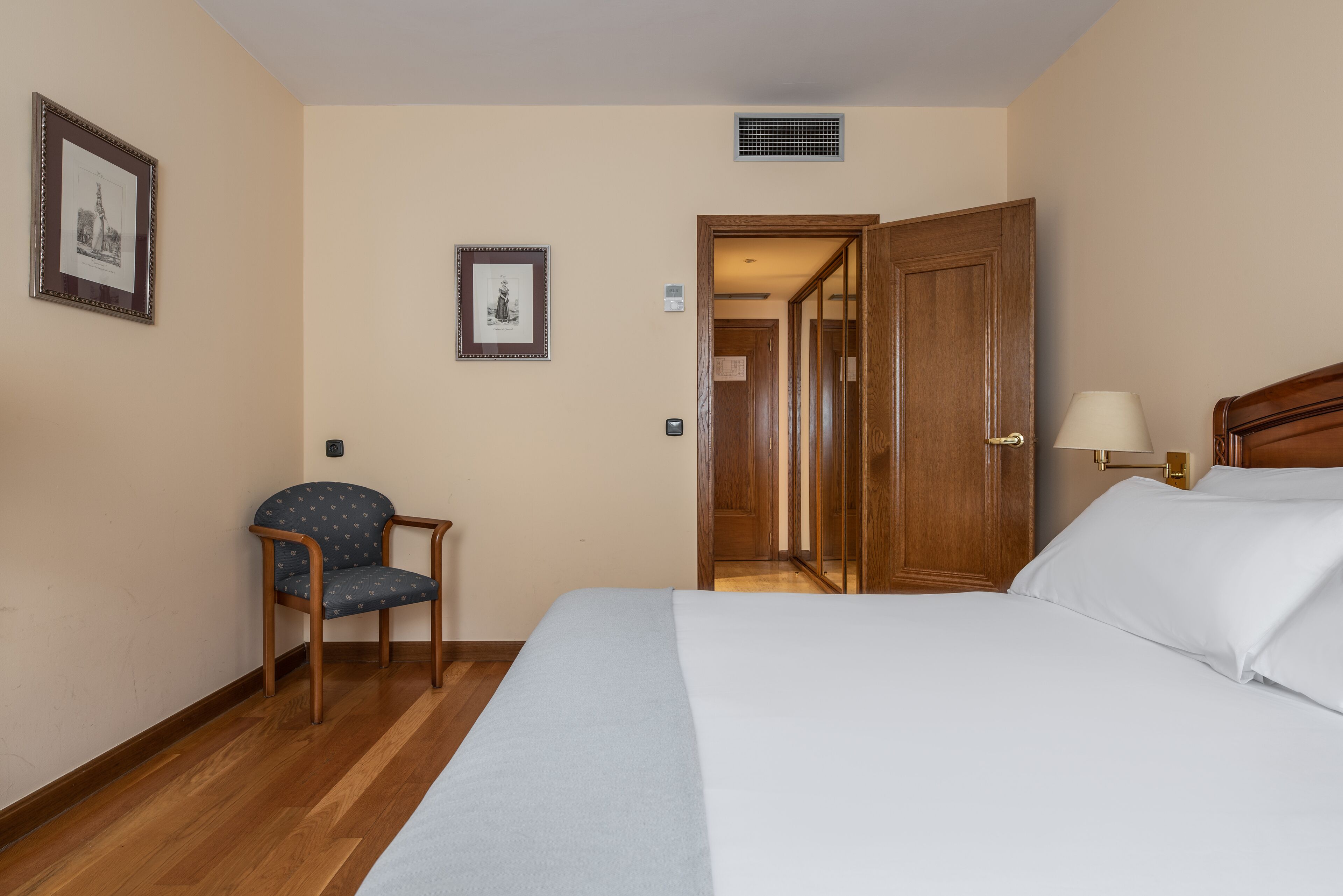 A hotel room with a bed, a chair, a lamp, and a door leading to another room.