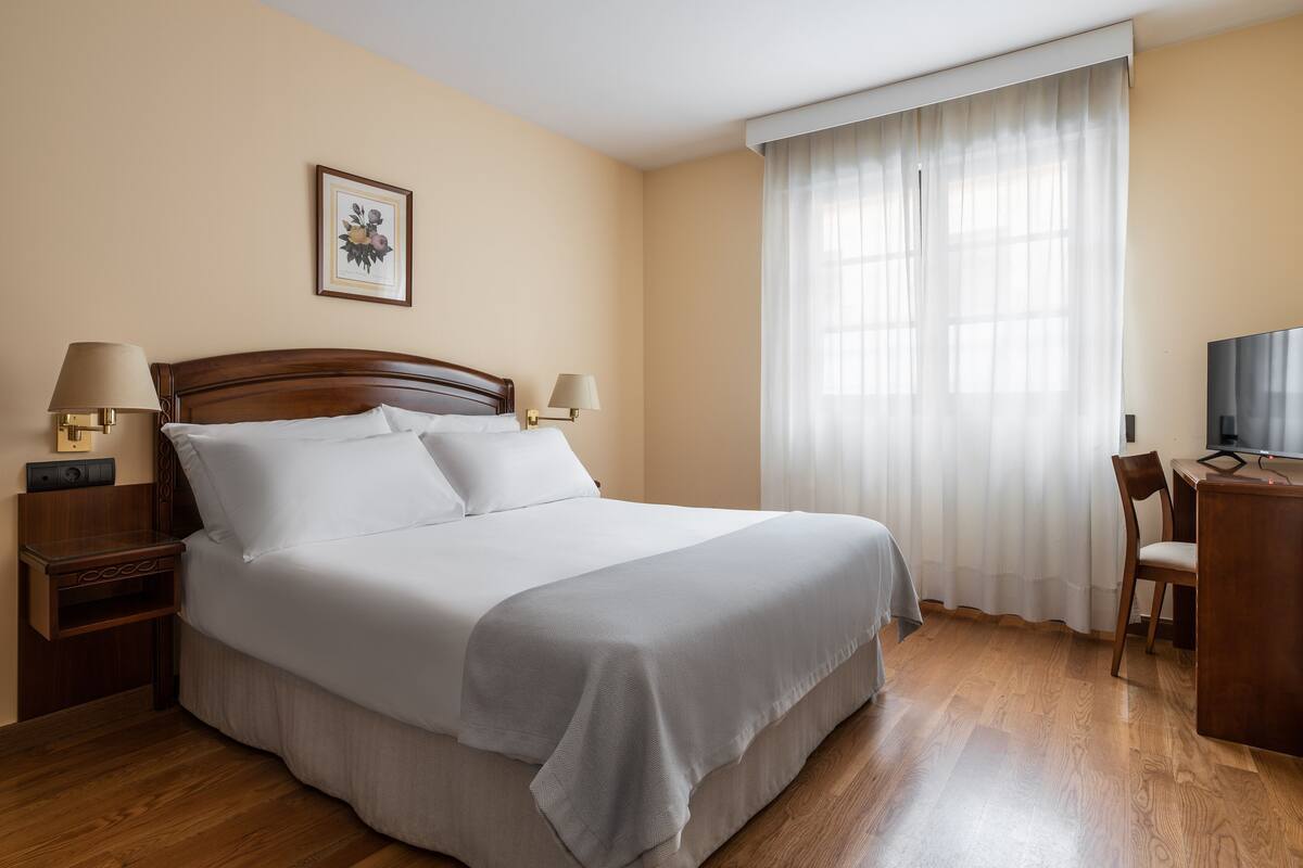 A hotel room with a large bed, two bedside tables with lamps, a desk with a chair, a TV, and a window with curtains.