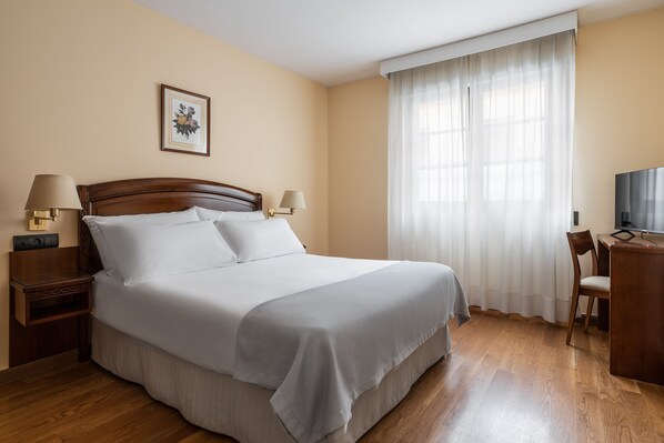 Habitación de hotel con una cama grande, dos mesitas de noche con lámparas, un escritorio con silla, un televisor y una ventana con cortinas.