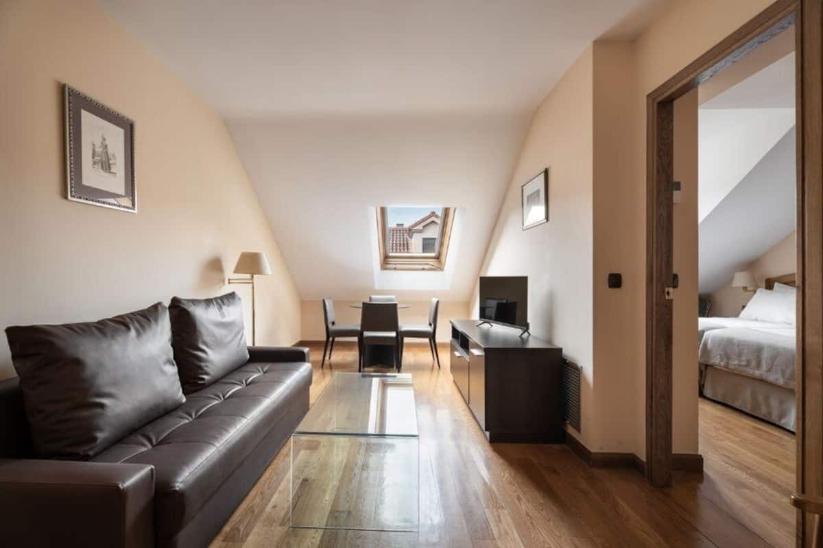 A modern, well-lit room with a brown leather sofa, a glass coffee table, a desk with a computer, and a bed.