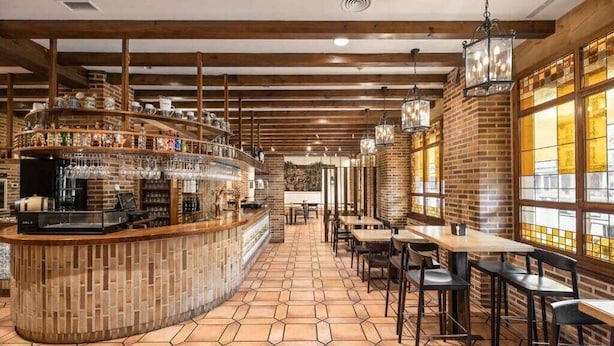 Zona de bar con mostrador, estanterías llenas de botellas e iluminación colgante. Hay mesas y sillas para sentarse.