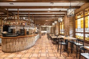 Un espace bar avec un comptoir, des étagères remplies de bouteilles et des luminaires suspendus. Des tables et des chaises sont à disposition pour s’asseoir.