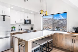 A modern kitchen with white cabinets, a stainless steel refrigerator, a microwave, and a large window offering a view of a snowy landscape. - Slopeside Escape w/ Pool, Hot Tubs & Views (Park City)