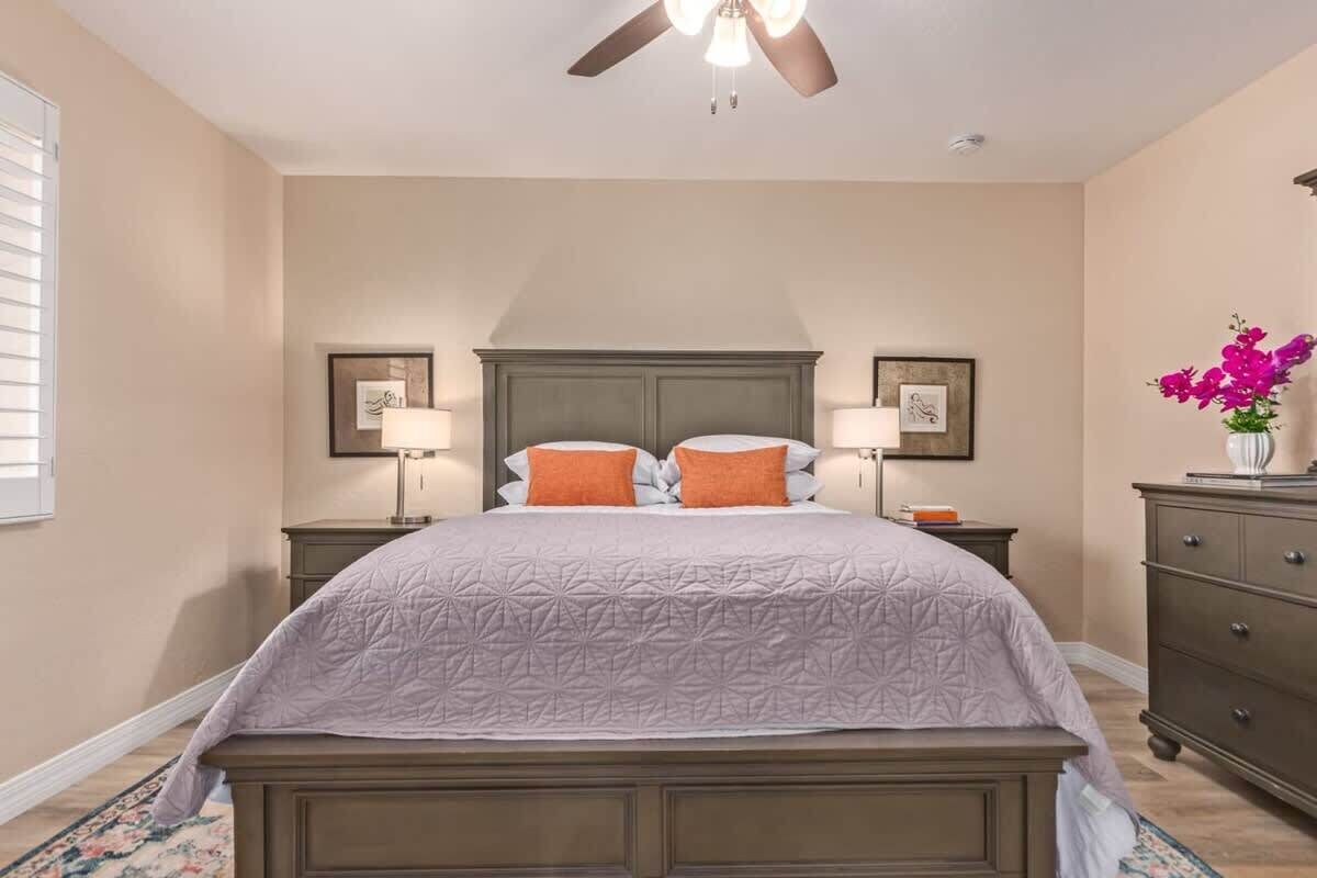 A bedroom with a bed, two bedside tables, a dresser, a ceiling fan, and a window with blinds.