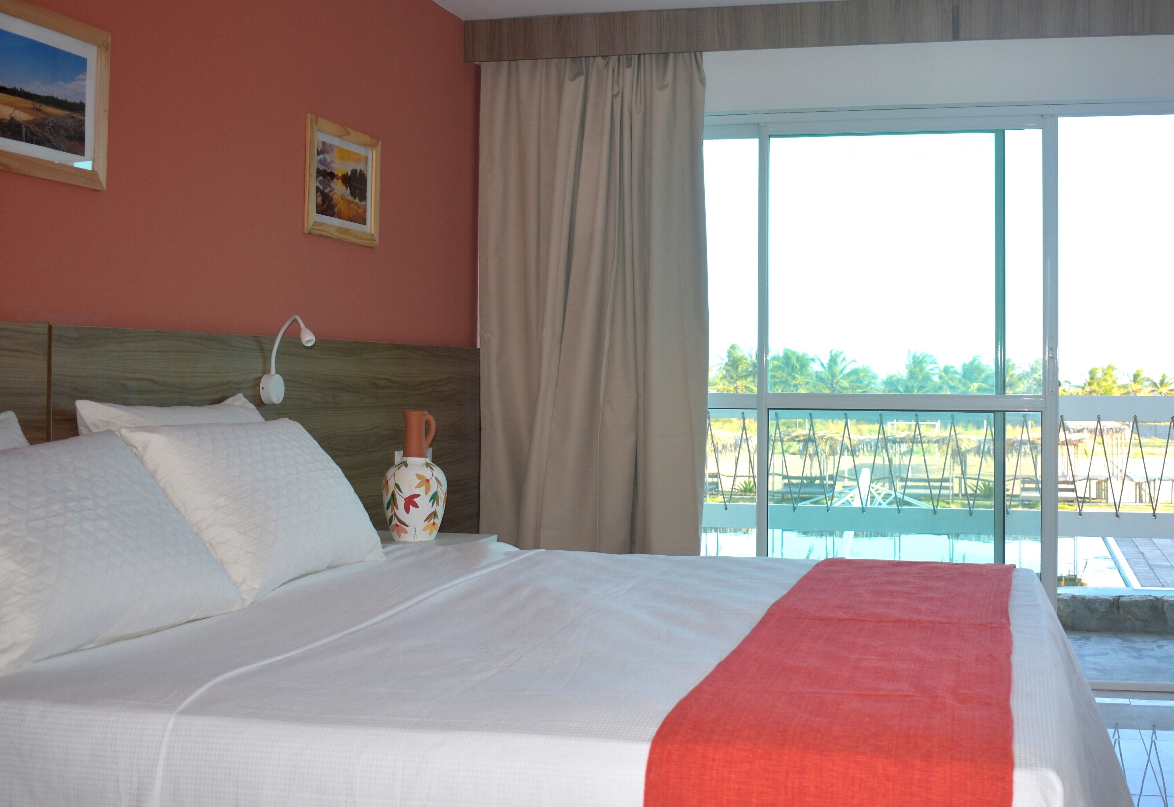 A hotel room with a large bed, a nightstand, a lamp, and a view of the pool and greenery outside.