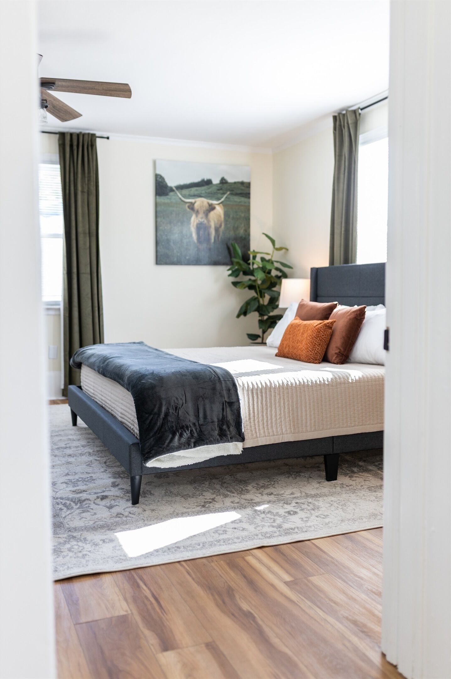A bedroom with a bed, a headboard, a painting, curtains, and a plant.