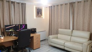 A room with a desk, two monitors, a printer, a black office chair, a beige sofa, and a radiator.