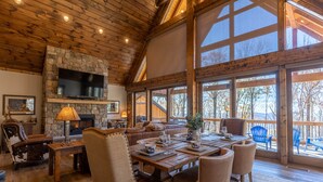 Un salón amplio con chimenea de piedra, techo de madera y ventanas grandes que ofrecen vistas al exterior.