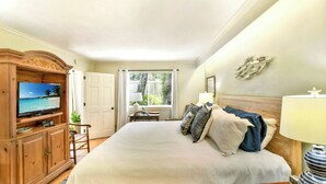 Ein Hotelzimmer mit einem großen Bett, einem Fernseher auf einer Holzkommode, einem Schreibtisch mit Stuhl und einem Fenster mit Jalousien.