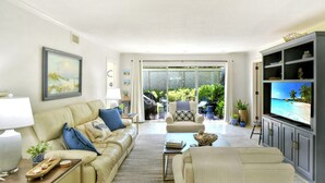 Ein Wohnzimmer mit beiger Couch, Couchtisch und Fernseher. Ein Schiebetürfenster führt in einen Außenbereich mit Pflanzen und Grill.