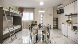 Een compacte kamer met een stapelbed, een eettafel met stoelen, een kitchenette met een magnetron en een aan de muur bevestigde televisie.