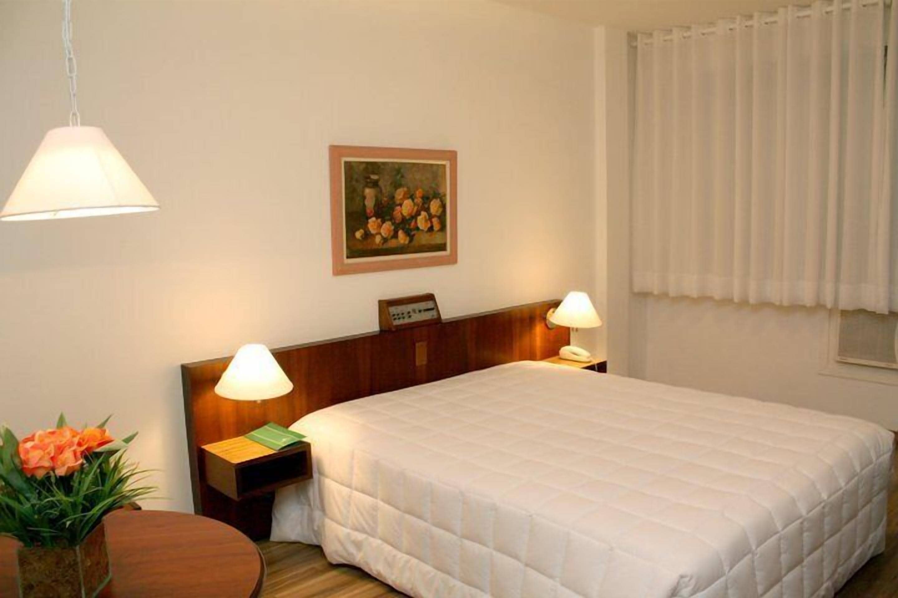 Quarto de hotel com uma cama grande, duas luminárias de cabeceira, uma mesa de cabeceira com abajur, um quadro na parede e uma janela com cortinas.